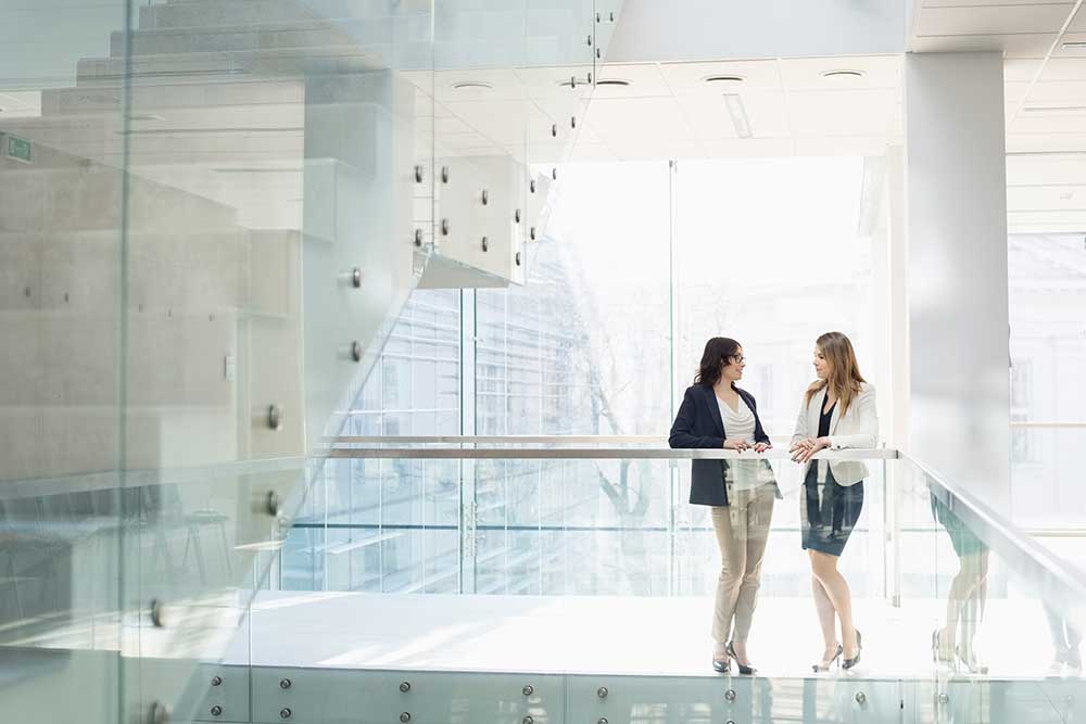 Two women leaning against the glass balustrade in a light-flooded stairwell.